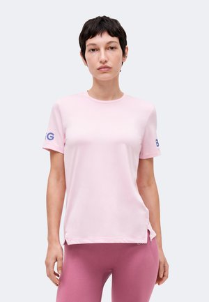 Kvinde med kort mørkt hår iført en lyserød kortærmet skjorte og mørkere lyserøde leggings, stående mod en ensfarvet baggrund.