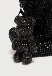 Mustast karutõllust võlu, kaunistatud peenete mustade kristallidega, kinnitatud musta koti külge, millel on detailne tekstuur ja läikiv viimistlus.