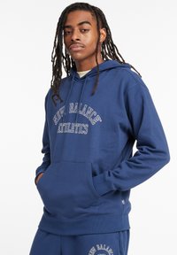 Giovane con lunghe trecce indossa una felpa blu New Balance Athletics e pantaloni coordinati, in piedi con le mani nelle tasche della felpa.