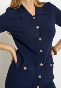 Marineblå strikket cardigan med krave, der har knapper i guldfarve og to forlommer, hvilket giver en tætsiddende silhuet.