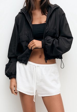 Vrouw die een zwarte cropped hooded jas draagt over een zwarte tube top en witte short met koord, staand tegen een witte achtergrond.
