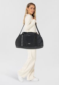 Borsa a mano nera in tessuto testurizzato, con manici doppi, logo e tasca con zip. Forma ovale elegante. Il modello indossa un top e pantaloni color crema.
