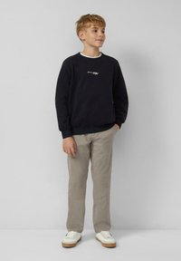 Ragazzo in piedi con le mani in tasca, indossa una felpa nera, pantaloni beigi e sneakers bianche, su uno sfondo chiaro e semplice.