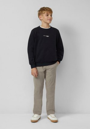 Junge steht mit Händen in den Taschen, trägt einen schwarzen Sweatshirt, beige Hosen und weiße Sneaker vor einem einfarbigen hellen Hintergrund.