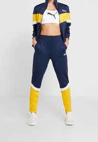 Marinblå idrottsjacka med gula och vita ränder, i kombination med matchande marinblå och gula joggerbyxor samt vita sneakers.