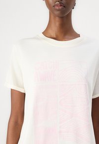Cremfarbener T-Shirt mit hochgerollten Ärmeln, mit pinkem grafiktext "CATCH A WAVE" und geometrischen Mustern auf der Vorderseite.