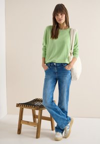 Femme portant un pull vert, un jean bleu, des baskets blanches et un sac à bandoulière blanc, debout à côté d'un tabouret en bois avec un siège tressé noir.