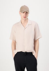 Camisa de botão de mangas curtas rosa claro com colarinho descontraído e um par de calças pretas. Inclui um boné bege e um brinco.