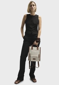 Sort ærmeløs top med knapper, tætsiddende sorte bukser og brune remsandaler. Beige tote taske med brune læderdetaljer og lynlåse.