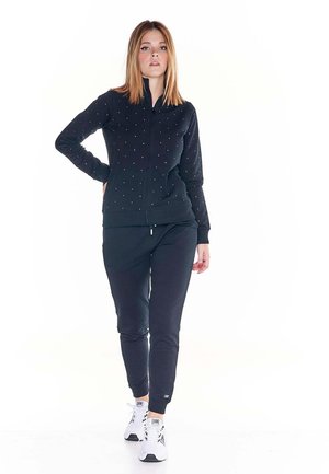 Une femme porte une veste noire zippée ornée de clous, un pantalon de jogging assorti et des baskets blanches, sur un fond blanc uni.