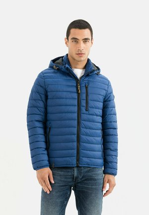 Polstret blå jakke med lynlås og hætte, der har lodrette striber, guld hardware-detaljer og sidelommer. Båret med jeans.