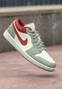 Sneaker basse avec des panneaux blancs, verts et rouges, lacets verts et un swoosh Nike rouge, flottant au-dessus d'une surface texturée rose.