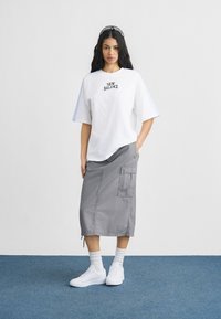 Λευκό βαμβακερό μπλουζάκι με το λογότυπο "NEW BALANCE", γκρι φούστα μέχρι το γόνατο με τσέπες και λευκά αθλητικά παπούτσια με ριγωτές κάλτσες μέχρι τον αστράγαλο.