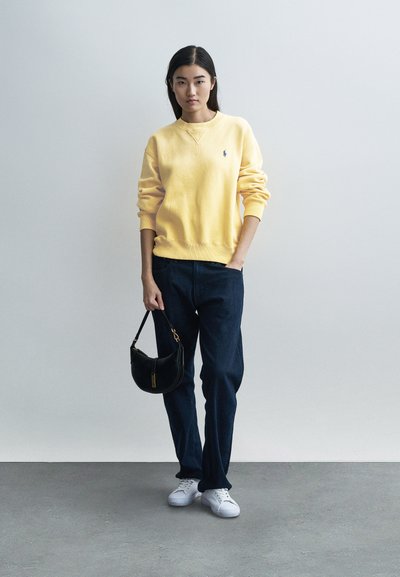 Yellow sweatshirt with a small logo, paired with dark blue jeans and white sneakers. Black handbag with gold hardware detail.