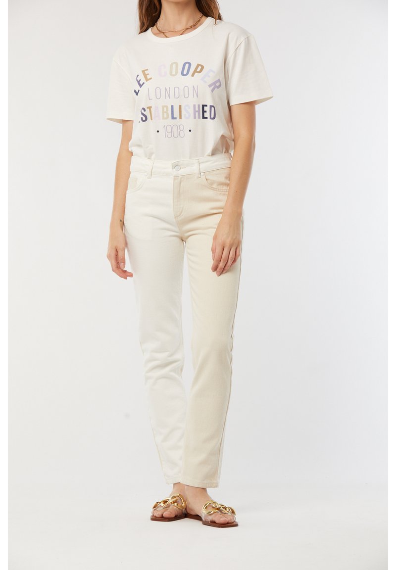 T-shirt en coton blanc avec un texte multicolore indiquant "Lee Cooper London Established 1908." Associé à un jean ajusté blanc cassé et des sandales accentuées de doré.