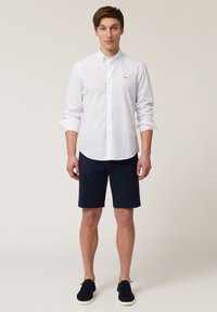 Camisa blanca de botones con patrones en azul claro, combinada con pantalones cortos navy y zapatos oscuros. La camisa tiene un cuello estándar y mangas largas.