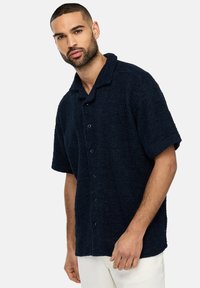 Camisa de manga corta azul marino hecha de tejido texturizado, con botones en la parte delantera y un cuello clásico. Textura notablemente suave y aterciopelada.