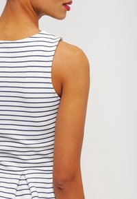 Halenka bez rukávů v bílo-námořnických pruzích, s vysokým výstřihem a drobným volánkem vzadu, vyrobena z hladkého materiálu.