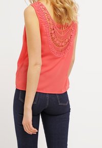 Blusa senza maniche di colore corallo con un design in pizzo sul retro. Tessuto morbido, scollatura tonda e vestibilità leggermente ampia. Abbinata a jeans in denim scuro.