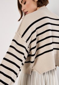 Pull en beige avec des rayures noires horizontales, texture côtelée, design court et détails d'épaules tombantes, superposé sur une légère jupe plissée.