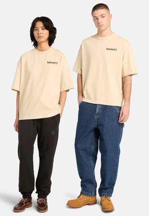 Dwójka modeli stoi przodem, ubrana w beżowe koszulki Timberland; jeden ma czarne spodnie dresowe i brązowe buty, drugi niebieskie dżinsy i jasnobrązowe botki.