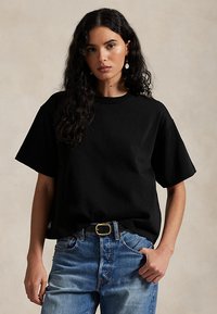 Žena v čiernom oversized tričku, modrých džínsoch s čiernym opaskom, stojí s jednou rukou v vrecku pred jednoduchým pozadím.