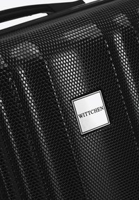 Valise rigide noire avec une texture à motif hexagonal, dotée de quincaillerie en argent et d'un petit logo de marque rectangulaire.