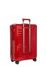 Valigia rigida rossa con superficie testurizzata e scanalature verticali, manico estraibile argento e quattro ruote nere.