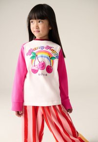 Pikkade varrukatega särk, millel on roosad varrukad ja valge kere, millel on värvikas kirsigraafika ja tekst. Triibulised püksid punaste ja roosade toonidega.