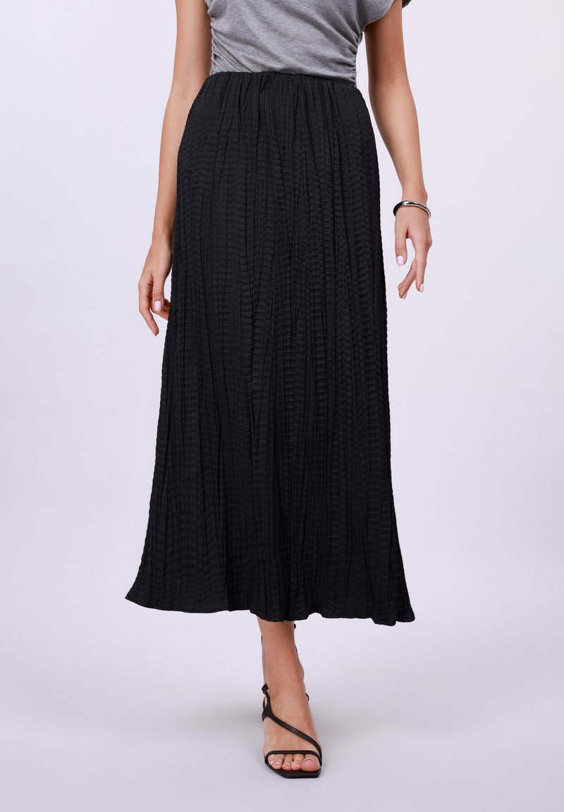 Zwarte plissé maxi-rok met een lichte textuur en een elastische tailleband, aangevuld met zwarte sandalen met bandjes.