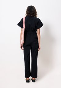 Zwart jumpsuit met korte mouwen, een getailleerde taille en wijde pijpen. Roze gestructureerde schoudertas met een geweven ontwerp. Zwarte plateauschoenen.