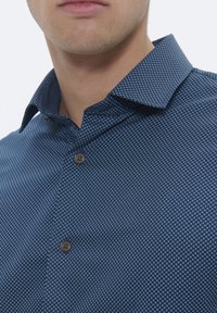 Camicia blu con motivo a miniature cerchi bianchi, colletto button-down, tessuto a trame e un singolo bottone decorativo sul davanti.