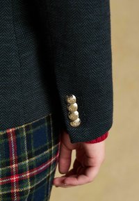 Tmavozelená herringbonová bunda má na rukáve štyri zlaté gombíky s kvetinovými vygravírovanými vzormi, v kombinácii s prekryvajúcimi sa kockovanými nohavicami.