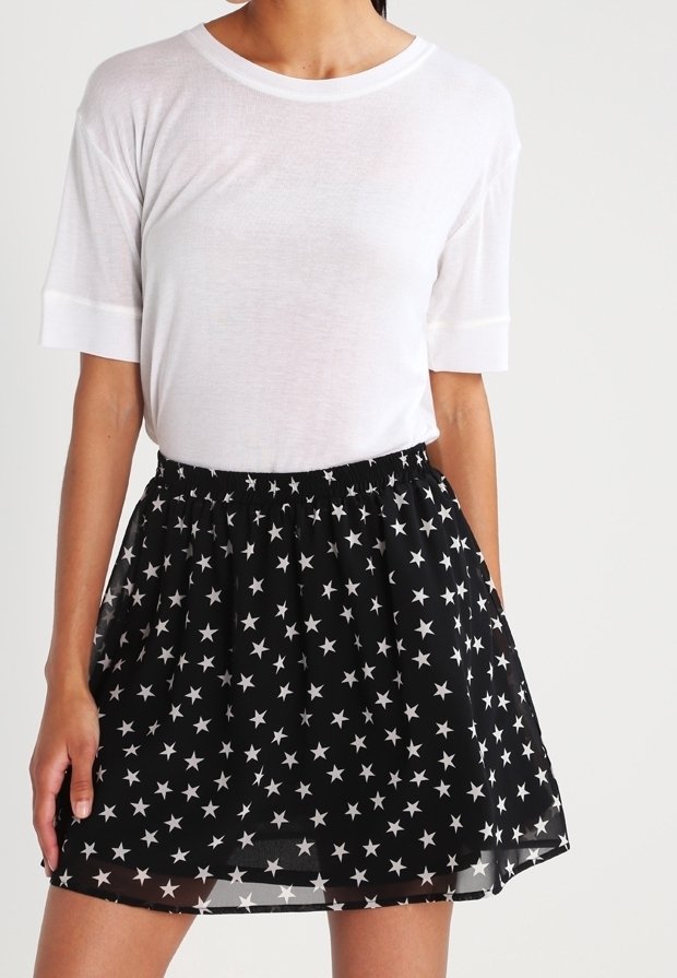 Femme portant une chemise blanche à manches courtes et une jupe noire avec des motifs d'étoiles blanches, debout devant un fond uni.