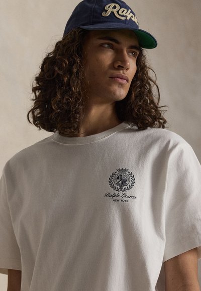 Junger Mann mit lockigem Haar, der ein weißes Ralph Lauren T-Shirt und eine marineblaue Kappe trägt, blickt seitlich vor einem schlichten Hintergrund.