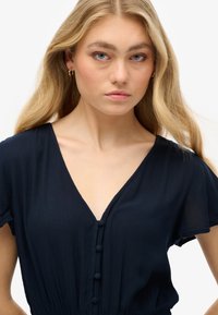 Mørkeblå, struktureret kjole med dyb V-hals, korte ærmer og knapdetaljer nedadtil. Glat stof med let drapering.