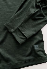 Chemise à manches longues vert foncé avec des coutures visibles et un petit logo triangulaire près de l'ourlet inférieur sur fond blanc.