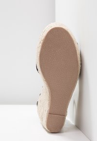 Talons compensés en espadrille avec une tige en jute, des accents en tissu noir et une semelle en caoutchouc texturé, mettant en valeur des matériaux naturels et une forme incurvée.