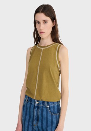 Mladá žena oblečená v olivovo zelenej tielkovej blúzke s kontrastnými švami a modrých pásikavých džínsoch, stojaca na jednoduchom pozadí.