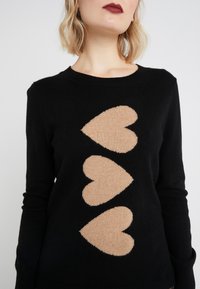 Frau in einem schwarzen Pullover mit drei großen beigen Herzen in vertikaler Reihe auf der Vorderseite, mit kurzem blonden Haar und dunklem Lippenstift.