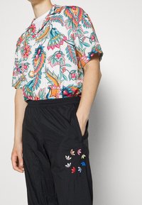 Lyhythihainen kukkakuosisel shirt, jossa on kirkkaat värit ja kuviot, yhdistettynä mustiin joustavaan vyötäröhousuihin, joissa on monivärisiä logotäydennyksiä.