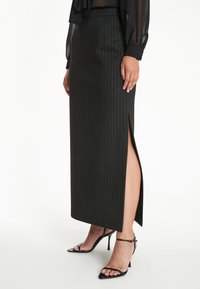 Zwarte maxi rok met streepjes en een zijsplit, gemaakt van een soepele stof. Gecombineerd met zwarte sandalen met hoge hakken en bandjes.