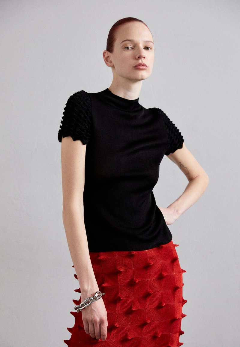 Top de punto negro con mangas abullonadas texturizadas; combinado con una falda roja con acentos moldeados en 3D. Una pulsera de plata gruesa adorna la muñeca.