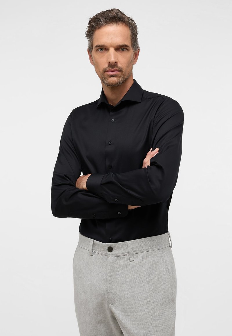 Camicia nera da uomo con bottoni e una texture liscia, maniche lunghe e colletto classico, abbinata a pantaloni grigi chiari con trama.