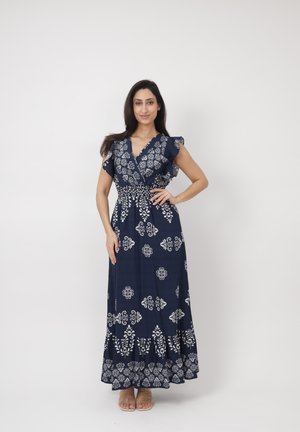 Femme debout, main sur la hanche, portant une robe longue bleu marine à motifs floraux blancs et manches flottantes, sur un fond clair uni.