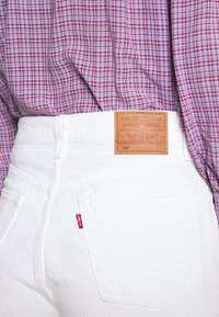 Jeans de denim blanco con una etiqueta de cuero marrón que dice "Levi Strauss & Co." y costuras rojas. La tapeta de una camisa de cuadros rojos y morados es visible.