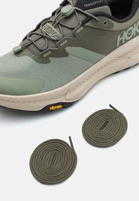 Zapatilla de correr HOKA verde con un upper texturizado, suela beige y logo Vibram negro. Dos cordones verdes enrollados al lado de la zapatilla.