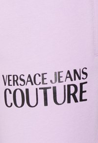 Levandulová látka s textem "VERSACE JEANS COUTURE" v černé barvě, s lesklým povrchem. Měkká textura a ležérní styl.