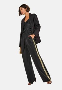 Cămașă de satin neagră asortată cu un sacou negru și pantaloni cu picioare largi, având o dungă laterală din paiete aurii. Modelul este pozat pe un fundal neutru.