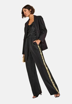 Camisole en satin noir associé à un blazer noir et un pantalon large avec une bande latérale en sequins dorés. Modèle posé contre un fond neutre.